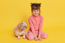 Girl With Puppy Isolated Over Yellow Background, Child Looking Directly At Camera With Smile, Kid Wearing Rose Sweater, Pants And Hair Band With Cat's Ears, Small Kid With Pekingese.