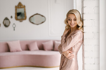 Attractive young blonde woman with curly hair and makeup in a sequined dress in the bright interior of the studio house