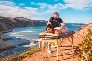 A young man performing a massage in nature on the coast near the sea, a dream come true, masseuse with face mask in the coronavirus pandemic