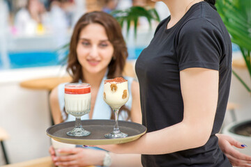 A young waitress carrying a tray with two different desserts in a two glass on the background of a young female client