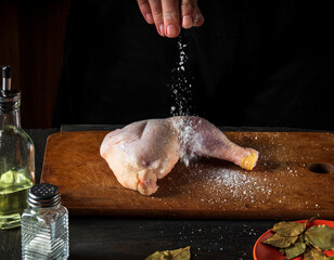 The chef sprinkles fresh raw chicken drumstick with sea salt on a dark background. There are ingredients and spices for cooking nearby