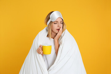 Wall Mural - Young woman in sleeping mask wrapped with blanket holding cup on yellow background