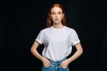 Wall Mural - Serious young woman inT-shirt and denim pants posing with hands in pockets on isolated black background, looking at camera. Pretty redhead lady model emotionally showing facial expressions,.