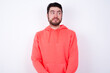 © Jihan - young Caucasian bearded man wearing pink hoodie against white background with thoughtful expression, looks away keeps hands down bitting his lip thinks about something pleasant.