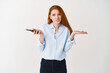 © Cookie Studio - Clueless young woman with red hair and blue shirt shrugging at camera, holding smartphone and looking confused, dont know, white background