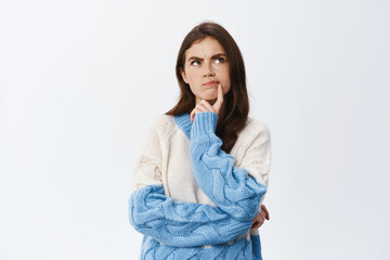 Pensive young woman having doubts, something off, frowning and touching lip while staring thoughtful at upper right corner, standing suspicious against white background