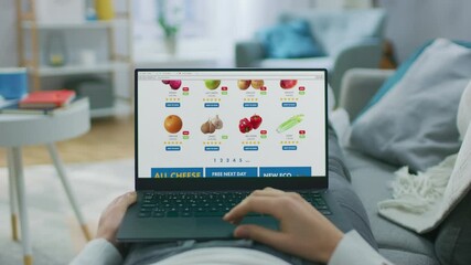 Wall Mural - Person Lying on Couch Uses Laptop Computer Browsing through Online Food Delivery Market, Choosing Through Vegetables Section, Fresh Goods. Internet Store Software with List of Food to Order. POV Shot