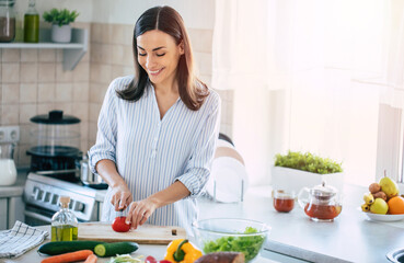 Wall Mural - Happy smiling cute woman is preparing a fresh healthy vegan salad with many vegetables in the kitchen at home and trying a new recipe