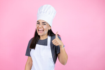 Wall Mural - Young woman wearing baker uniform with flour on the face over pink background success sign doing positive gesture with hand, thumb up smiling and happy. cheerful expression and winner gesture.