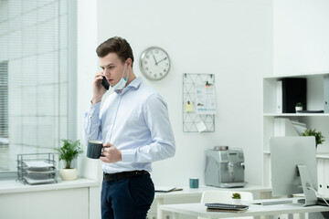 Poster - Young male office worker with cup of hot drink consulting client on smartphone