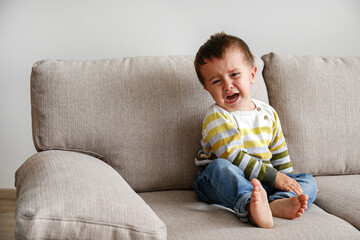 Portrait of adorable little boy sitting on the textile couch and crying. Upset toddler throwing a tantrum at home. Barefoot kid calling for attention. Close up, copy space, background.