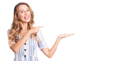 Young caucasian woman with blond hair wearing summer dress amazed and smiling to the camera while presenting with hand and pointing with finger.