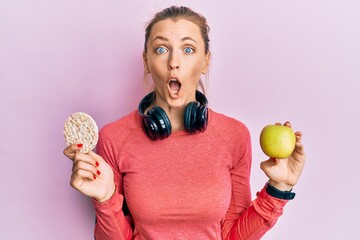 Sticker - Beautiful caucasian sports woman holding green apple and rice crackers afraid and shocked with surprise and amazed expression, fear and excited face.