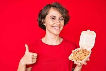 Poster - Young hispanic woman holding potato chip smiling happy and positive, thumb up doing excellent and approval sign