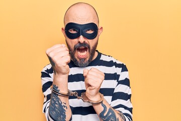 Sticker - Young handsome man wearing burglar mask and handcuffs annoyed and frustrated shouting with anger, yelling crazy with anger and hand raised