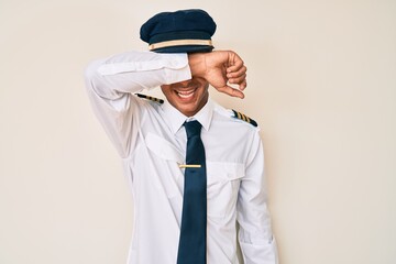 Sticker - Young hispanic man wearing airplane pilot uniform covering eyes with arm smiling cheerful and funny. blind concept.