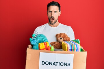 Sticker - Young handsome man holding donation box with toys in shock face, looking skeptical and sarcastic, surprised with open mouth