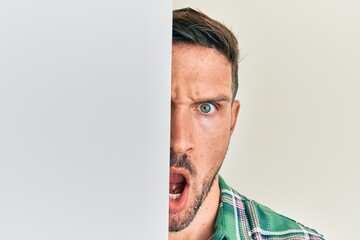 Sticker - Handsome man with beard holding blank empty banner covering half face in shock face, looking skeptical and sarcastic, surprised with open mouth