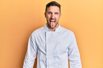 Canvas Print - Handsome man with beard wearing professional cook uniform sticking tongue out happy with funny expression. emotion concept.