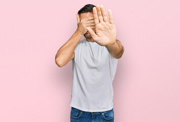 Wall Mural - Handsome man with beard wearing casual white t shirt covering eyes with hands and doing stop gesture with sad and fear expression. embarrassed and negative concept.