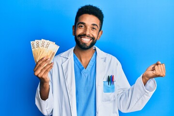 Poster - Handsome hispanic man with beard wearing medical uniform holding 500 norwegian krone screaming proud, celebrating victory and success very excited with raised arm