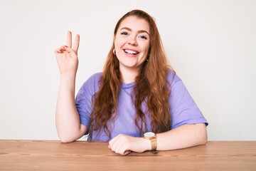 Poster - Young redhead woman wearing casual clothes sitting on the table smiling with happy face winking at the camera doing victory sign. number two.