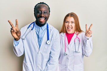 Sticker - Young interracial couple wearing doctor uniform and stethoscope smiling with happy face winking at the camera doing victory sign. number two.