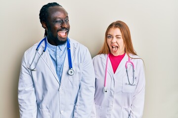 Wall Mural - Young interracial couple wearing doctor uniform and stethoscope winking looking at the camera with sexy expression, cheerful and happy face.