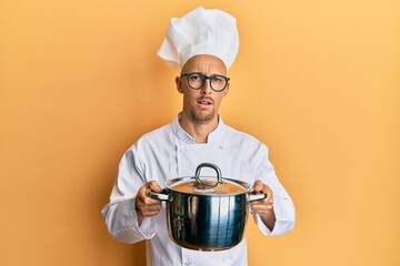 bald man with beard wearing professional cook holding cooking pot in shock face, looking skeptical a