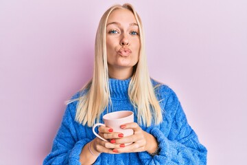 Poster - Young blonde girl wearing winter sweater and drinking a cup of hot coffee looking at the camera blowing a kiss being lovely and sexy. love expression.