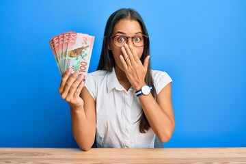 Sticker - Beautiful hispanic woman holding 100 new zealand dollars banknote covering mouth with hand, shocked and afraid for mistake. surprised expression