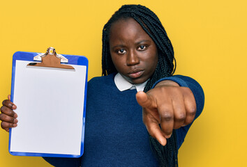 Sticker - Young black woman with braids holding clipboard with blank space pointing with finger to the camera and to you, confident gesture looking serious