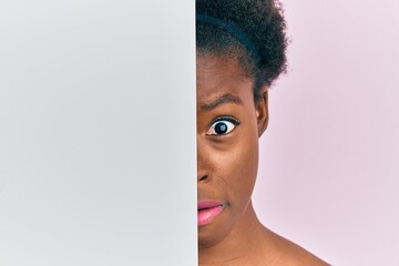 Canvas Print - Young african american girl holding blank empty banner over half face clueless and confused expression. doubt concept.