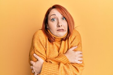 Poster - Beautiful redhead woman wearing casual winter sweater over yellow background shaking and freezing for winter cold with sad and shock expression on face