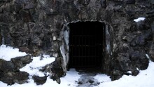 Old Stone Wall With Grid Door And Melting Snow. Entrance To Old Castle With Thick Stone Walls And Dark Room Inside The Castle. Red String Lights Visible Inside The Room.