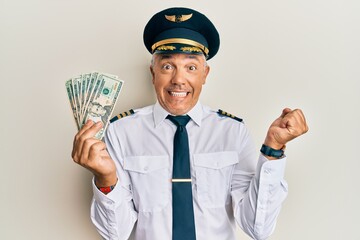 Canvas Print - Handsome middle age mature man wearing airplane pilot uniform holding dollars screaming proud, celebrating victory and success very excited with raised arm