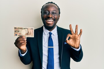 Sticker - Handsome young black man wearing business suit holding 1000 yen banknotes doing ok sign with fingers, smiling friendly gesturing excellent symbol