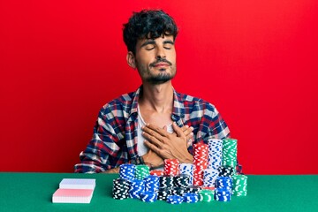 Sticker - Young hispanic man sitting on the table with poker chips and cards smiling with hands on chest, eyes closed with grateful gesture on face. health concept.