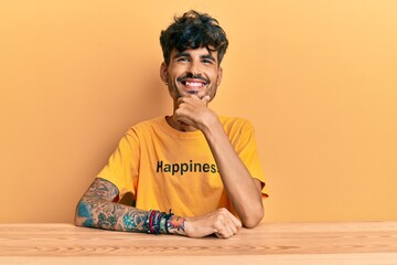 Wall Mural - Young hispanic man wearing tshirt with happiness word message sitting on the table looking confident at the camera smiling with crossed arms and hand raised on chin. thinking positive.