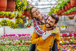 Smiling couple in love having piggyback ride in greenhouse. All around them are colorful flowers. Small business owners.