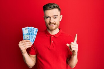 Wall Mural - Young redhead man holding 1000 hungarian forint banknotes smiling with an idea or question pointing finger with happy face, number one