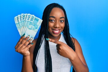 Young african american woman holding 100 brazilian real banknotes smiling happy pointing with hand and finger