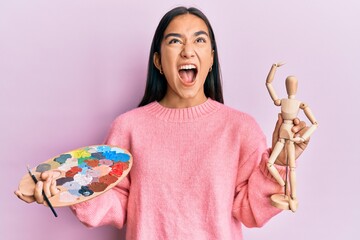 Canvas Print - Young asian woman holding painter palette and art manikin angry and mad screaming frustrated and furious, shouting with anger looking up.