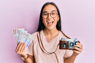 Sticker - Young asian woman holding vintage camera and swedish krona smiling and laughing hard out loud because funny crazy joke.