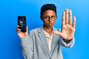 Wall Mural - Young african american girl holding broken smartphone showing cracked screen with open hand doing stop sign with serious and confident expression, defense gesture