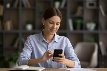 Canvas Print - Happy millennial Caucasian woman sit at desk at home office look at cellphone screen browse internet. Smiling young female use smartphone take notes. Communication, technology concept.