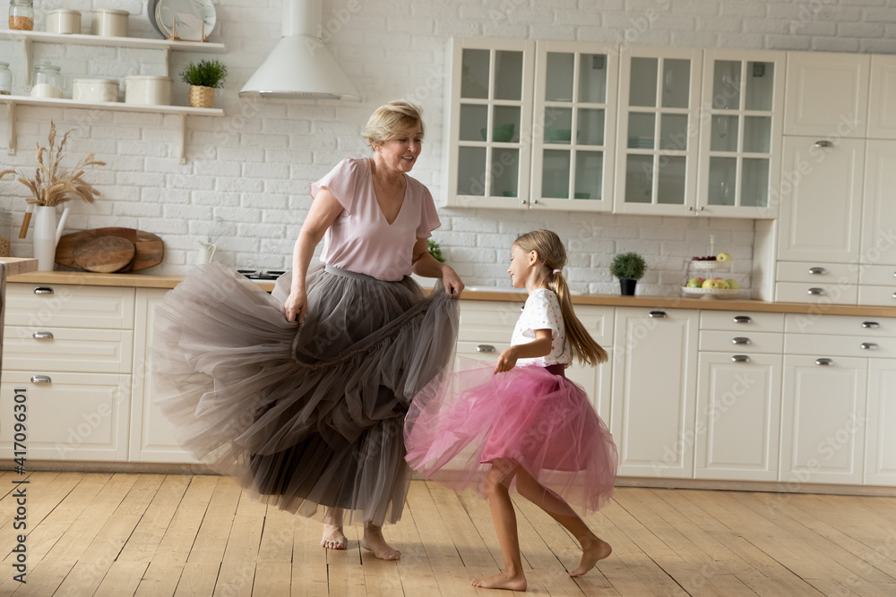 Merry leisure time. Happy energetic grandmother teach ball dance active little child. Caring grandma junior girl grandkid engaged in dancing in funny large fluffy skirts barefoot on warm kitchen floor - obrazy, fototapety, plakaty 