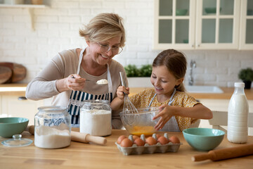 Cooking with soul. Happy little girl in apron help senior grandma at kitchen mix dough for cookies pancakes. Smiling older granny teach small grandkid to bake homemade cake pastry share family recipe