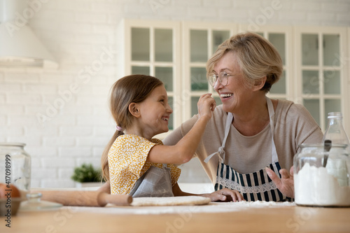 Funny baking. Joyful little girl having fun with retired older grandma when cooking pastries. Laughing elderly granny and cute small grandchild playing at kitchen painting one another noses with flour