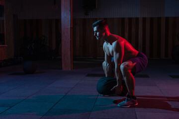 Wall Mural - A muscular powerful man with a naked torso exercises with a heavy bag in a red blue neon light. Cross workout in a modern gym